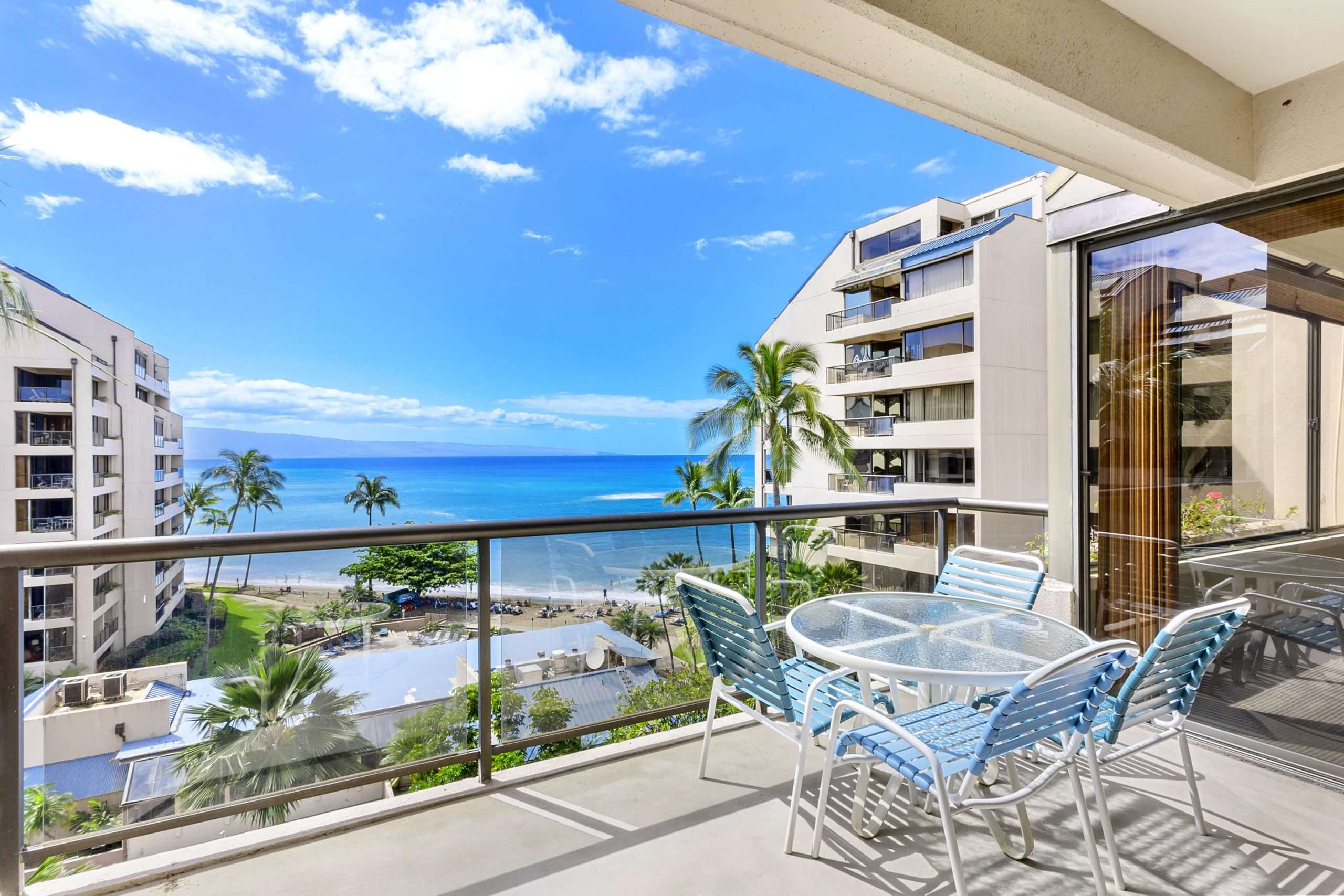 Ocean-view balcony with glass railing, table and chairs overlooking palm trees, pool deck, and the Pacific between resort towers.