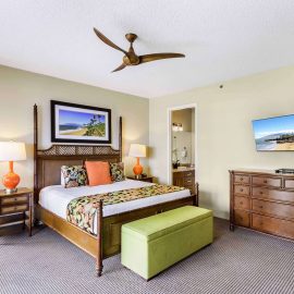 Spacious suite bedroom featuring a four-poster king bed, orange lamps, green bench, and dresser under a wall-mounted TV.