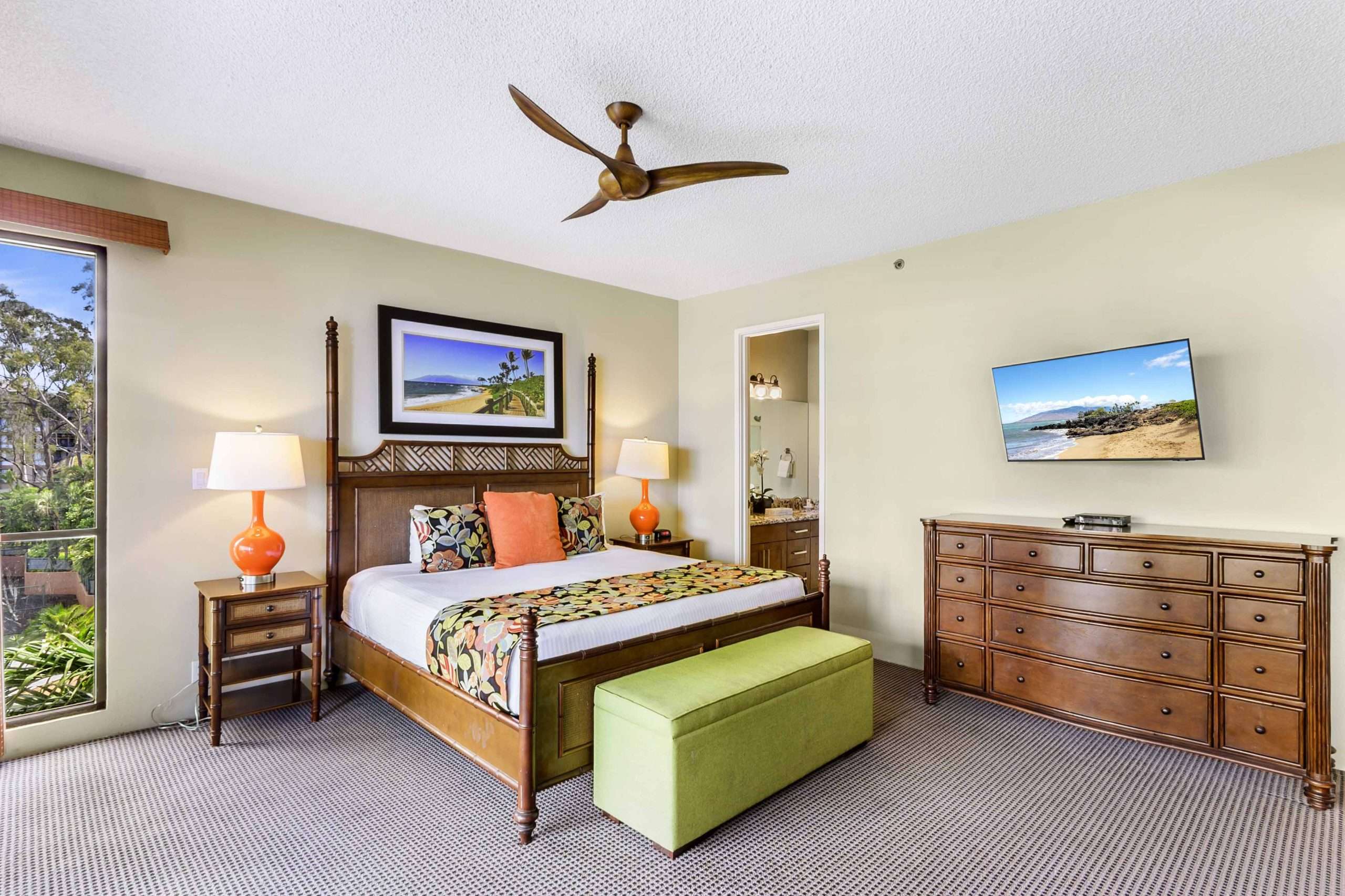 Spacious suite bedroom featuring a four-poster king bed, orange lamps, green bench, and dresser under a wall-mounted TV.