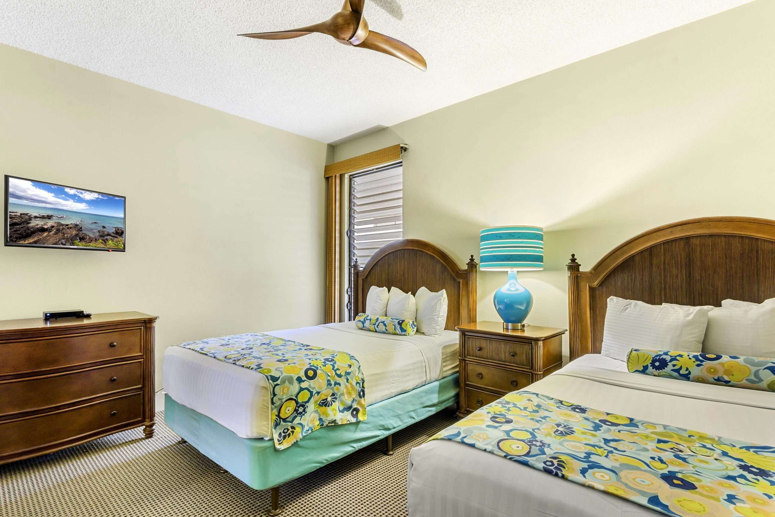Two queen beds with floral runners, bedside table and lamp, and wood dresser in a Maui two-bedroom suite.