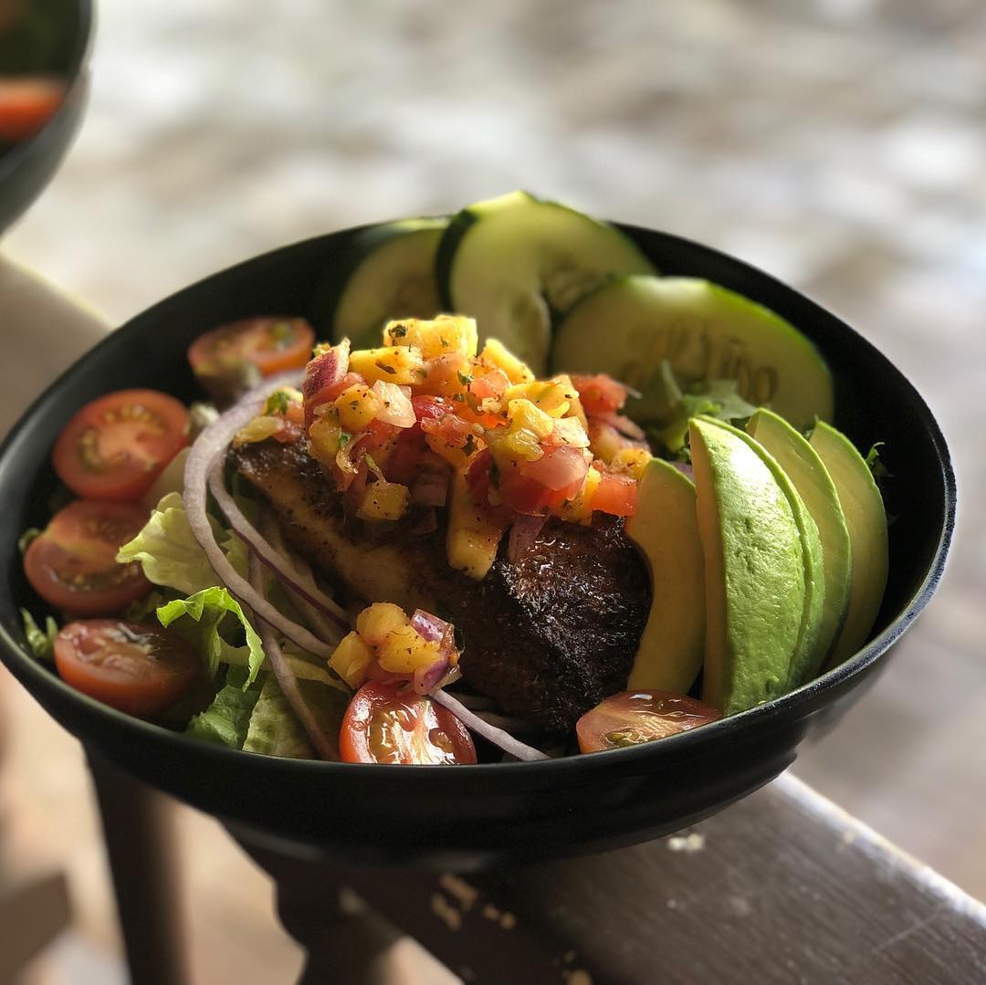 Grilled fish topped with pineapple salsa in a bowl of lettuce, avocado slices, cucumber, and cherry tomatoes.